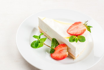 Light dessert: classic cheesecake, decorated with mint and strawberries. On a white plate, on a white wooden table. 