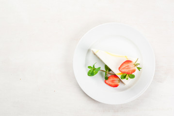 Light dessert: classic cheesecake, decorated with mint and strawberries. On a white plate, on a white wooden table. 