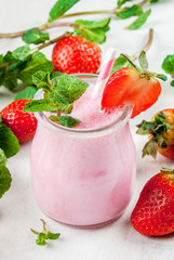Summer dessert or breakfast - strawberry milkshake with mint, in a small jar, with a striped tube. On a white table. copy space