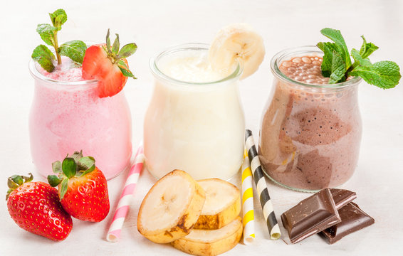 Selection of classic milkshakes in small jars: chocolate, strawberry, banana. On a white wooden table, with tubes, copy space