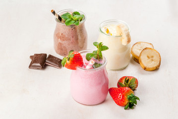 Selection of classic milkshakes in small jars: chocolate, strawberry, banana. On a white wooden table, with tubes, copy space