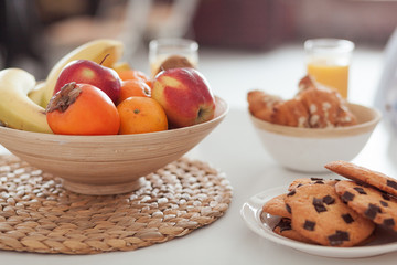 Plate of tasty cookies with chocolate crumbs, croissant, bananas, apples, persimmon, orange and orange juice on white table