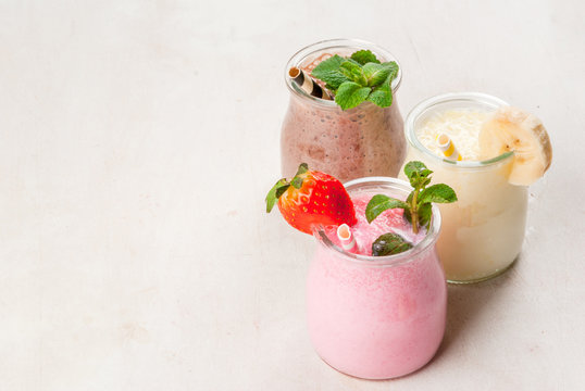 Selection of classic milkshakes in small jars: chocolate, strawberry, banana. On a white wooden table, with tubes, copy space