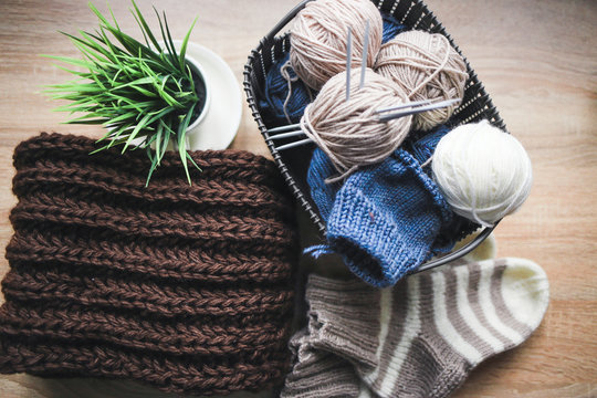 Beige, white and blue yarn, knitting needles in the basket and a brown scarf. Striped beige-white knitted socks and a green plant in the pot. Wooden background. Knitting hobby