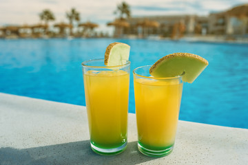 Glasses with yellow and green cocktail in front of a swimming pool with clear water. Close-up photo. Palm trees on the background.