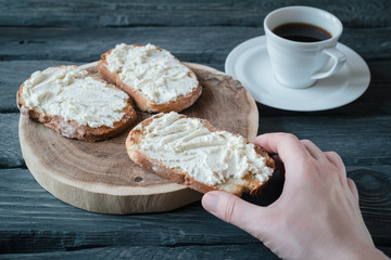 Hand holds homemade sandwiche with cream-cheese over a black wooden table. Wooden stand with sandwiches. Breakfast with black coffee.