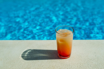 Glass with orange cocktail in front of a swimming pool with clear water. Close-up photo. Place for text