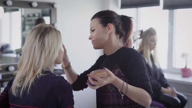 Beauty shop. The master in a make-up trains the student for final. The girl puts a brush foundation on a face of the client. The makeup artist professionally does a make-up.