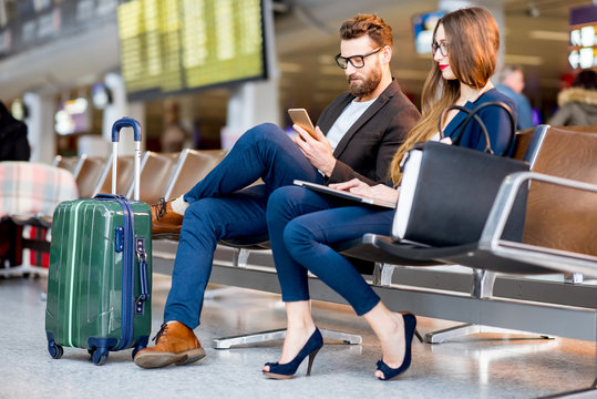 Elegant business couple sitting with phone and book at the waiting hall in the airport. Business travel concept