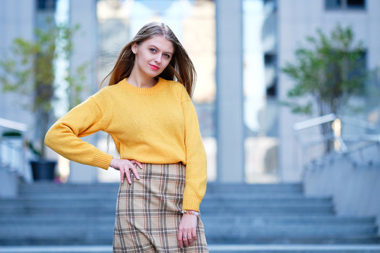 Blond hair young woman in yellow jamper and check skirt posing and against modern building. Street fashion style concept. 