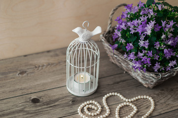 White candlestick in the form of cells with a bird. Wicker basket with violet flowers and beautiful white pearl necklace on a wooden background.