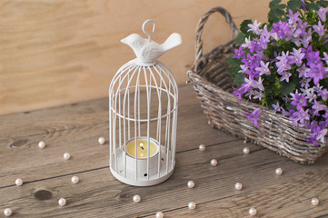 Little light gray wicker basket with beautiful Campanula portenschlagiana. White candlestick in the form of cells with a bird on the surface and scattered beads of pearls.