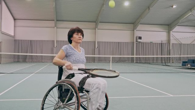 Disabled mature woman on wheelchair playing tennis on tennis court.