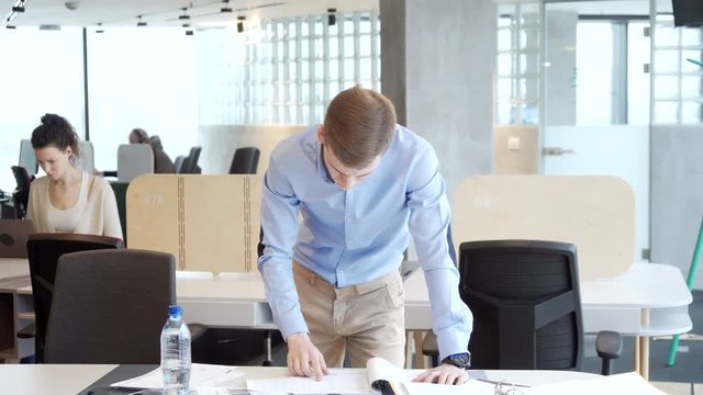 young working entrepreneur look for information among piles of papers, documents and folder for business analytics tasks staying at the table in the office open space during sunny day