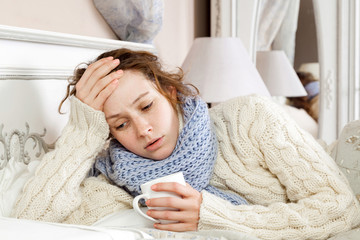 Sick woman with cup of tea. Closeup image of young frustrated sick woman in knitted blue scarf holding a cup of tea while lying in bed. hand on head, sad and feeling bad.