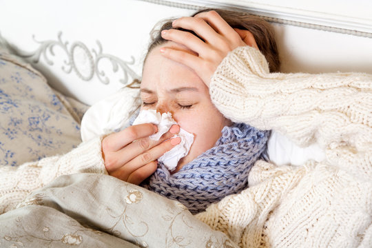 Flu. Closeup image of frustrated sick woman lying in bed in thick blue scarf holding tissue by her nose and touching her head and blowing her nose with closed eyes.
