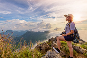 Hiker asian young woman happy with camera backpack and caps looking beautiful landscape nature of mountain and colourful sky at sunset on viewpoint Phu Chi Fa Forest Park, Chiang Rai, Thailand