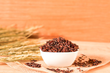 the brown raw rice in bowl with  dried rice plant on wooden table background.