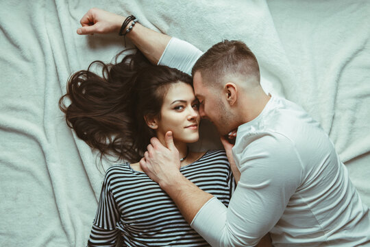 Portrait Of Beautiful Romantic Young Couple Man Woman In Love Hugging, Kissing, Lying On Floor On White Rug Carpet Indoors At Home, Toned With Filters, View From Top Above, Authentic Lifestyle