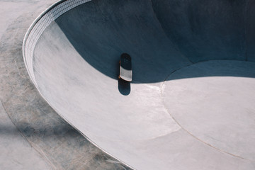 Skateboard lies on a concrete ground in a skate pool during daylight, with shadows and highlight on it