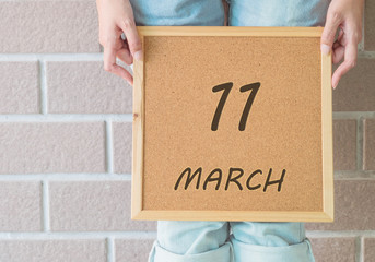 Closeup calendar at the cork board in hand of asian woman in front of her legs with 11 march word on brick wall textured background