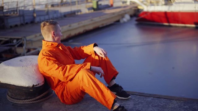 Young man in orange uniform sitting during his break by the sea in the harbor
