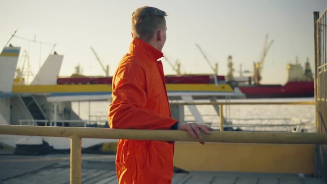 Young man in orange uniform standing by the fence and resting during his break by the sea in the harbor