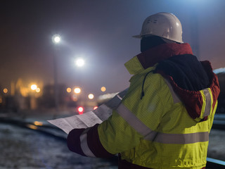 Back view of young construction worker in helmet at night reading construction drawings, prints.