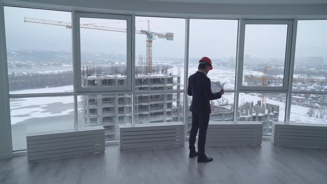 architect holding a design plan or other technical drawing looking on the project . worker in elegant suit and hard hat looking through the windows on the construction site with crane and workers on