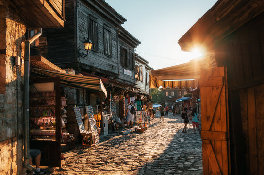 People walk through pedestrian cobblestone streets of ancient Nessebar with cafe, restaurant and souvenir shops at sunset.