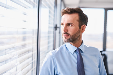 Portrait of handsome businessman standing at office looking out the window - Powered by Adobe