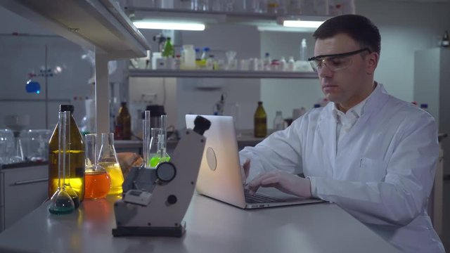Chemist Engineer working with laptop in laboratory. Adult man wearing in lab coat typing on computer. Caucasian professional entering data focused at work in the evening.