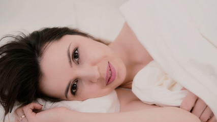 Obraz na płótnie Canvas Fresh and happy brunette woman in bed at home. Beautiful girl hides under a blanket and smiling.