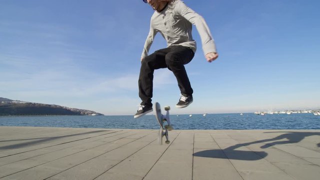 CLOSE UP SLOW MOTION: Young pro skateboarder skateboarding and jumping ollie flip trick on promenade along the coast in sunny summer. Skateboarder jumping kickflip trick with skateboard near the ocean