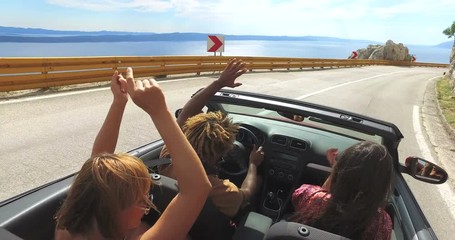 Friends dancing and waving arms driving on road along coast in red convertible