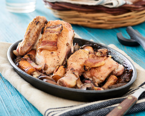 Baked chicken in beer sauce with mushroom, carrots and bacon in an iron cast baking pan on a blue table, with some bread slices in a basket