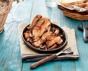 Baked chicken in beer sauce with mushroom, carrots and bacon in an iron cast baking pan on a blue table, with some bread slices in a basket