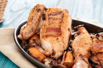 Baked chicken in beer sauce with mushroom, carrots and bacon in an iron cast baking pan on a blue table, with some bread slices in a basket