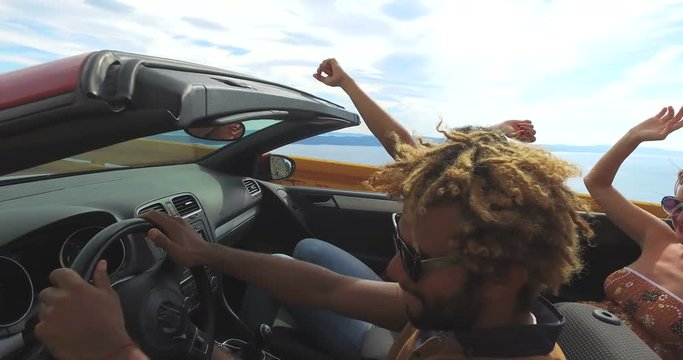 Side view of friends waving arms driving on coastal road in red convertible