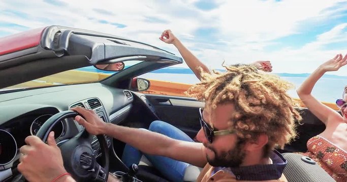 Side view of friends waving arms driving on coastal road in convertible, graded