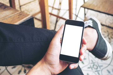 Mockup image of hand holding black mobile phone with blank white screen on thigh with black canvas shoes at vintage tile floor in cafe 