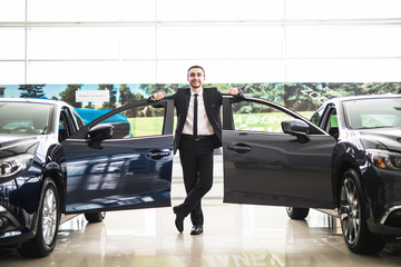 Confident smiling car salesman at the showroom near two cars, he is standing near open cars doors...