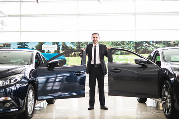 Confident smiling car salesman at the showroom near two cars, he is standing near open cars doors...