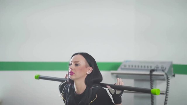 Striking Brunette holds bodibar shoulders and doing squats. Woman connected to sensors - EMS fitness. Electrical stimulation of muscles. Modern type of fitnes - EMS