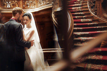 elegant gorgeous bride and stylish groom hugging, romantic moment. standing on wooden stairs in rich room. unusual wedding couple in retro style.