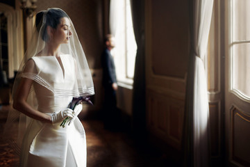 elegant gorgeous bride gently looking under veil at stylish groom standing at window light, holding...