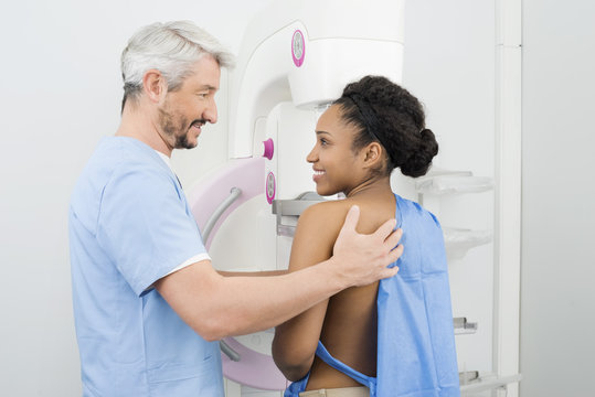 Smiling Doctor Looking At Female Patient Undergoing Mammogram Te