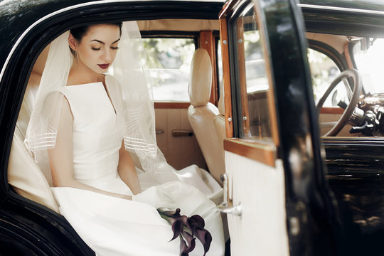 gorgeous elegant bride posing in stylish retro black car, sitting inside in saloon holding bouquet of callas. luxury wedding in vintage style. unusual gown and veil