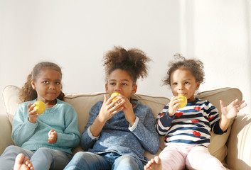 Three cute African girls sitting on sofa and eating lemons