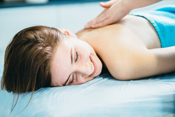 Close up smiling young woman lying vestured turquoise towel while massage therapist massaging her...